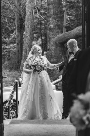 A bride in a lace gown holds a bouquet while interacting with someone on outdoor steps.