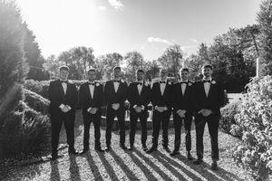 A group of groomsmen in black tuxedos stand in a line outdoors casting long shadows on a sunny day.