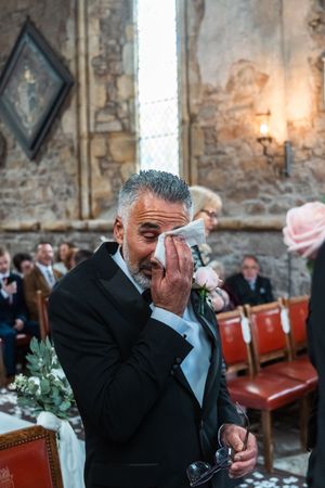 An emotional moment captured at a church wedding as a man in a black suit wipes tears from his eyes.