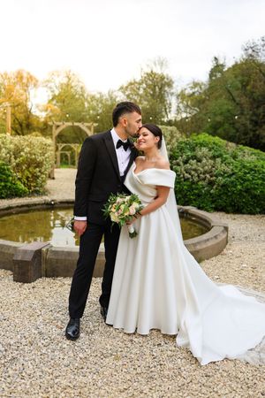 Romantic newlyweds share intimate moments on gravel path by garden pond during golden hour wedding photoshoot.