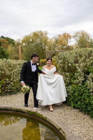 A couple in wedding attire shares a playful moment near a garden pond on their special day.