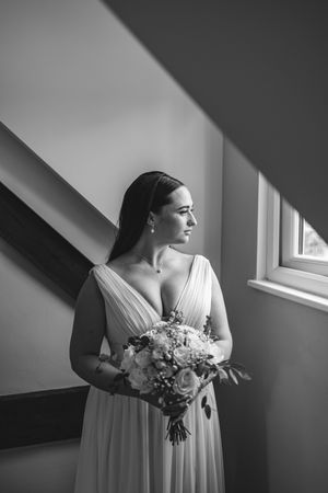 Black and white portrait of a bride in a flowing white dress holding a bouquet near a window.