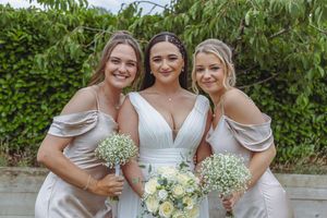 A group in coordinated light-colored dresses pose together at an outdoor wedding celebration with greenery in the background.