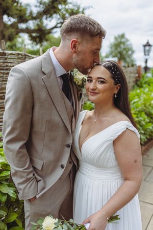 Romantic wedding couple in white dress and tan suit embracing outdoors on a sunny day.