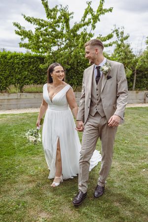 A couple in wedding attire walks hand in hand across a green lawn on their special day.