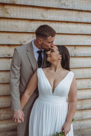 Newlyweds in white dress and grey suit share tender moment against rustic wooden wall backdrop.