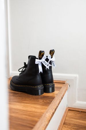 Black Dr. Martens combat boots with white bow detail sit on wooden stairs against a white wall.