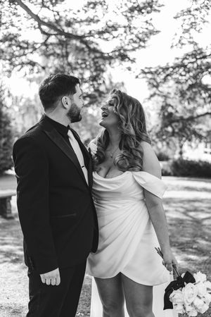 Black and white photo showing couple in wedding attire sharing intimate moment outdoors with trees in background.
