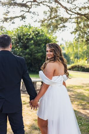 A couple in wedding attire holds hands while standing in a sunlit outdoor garden setting.