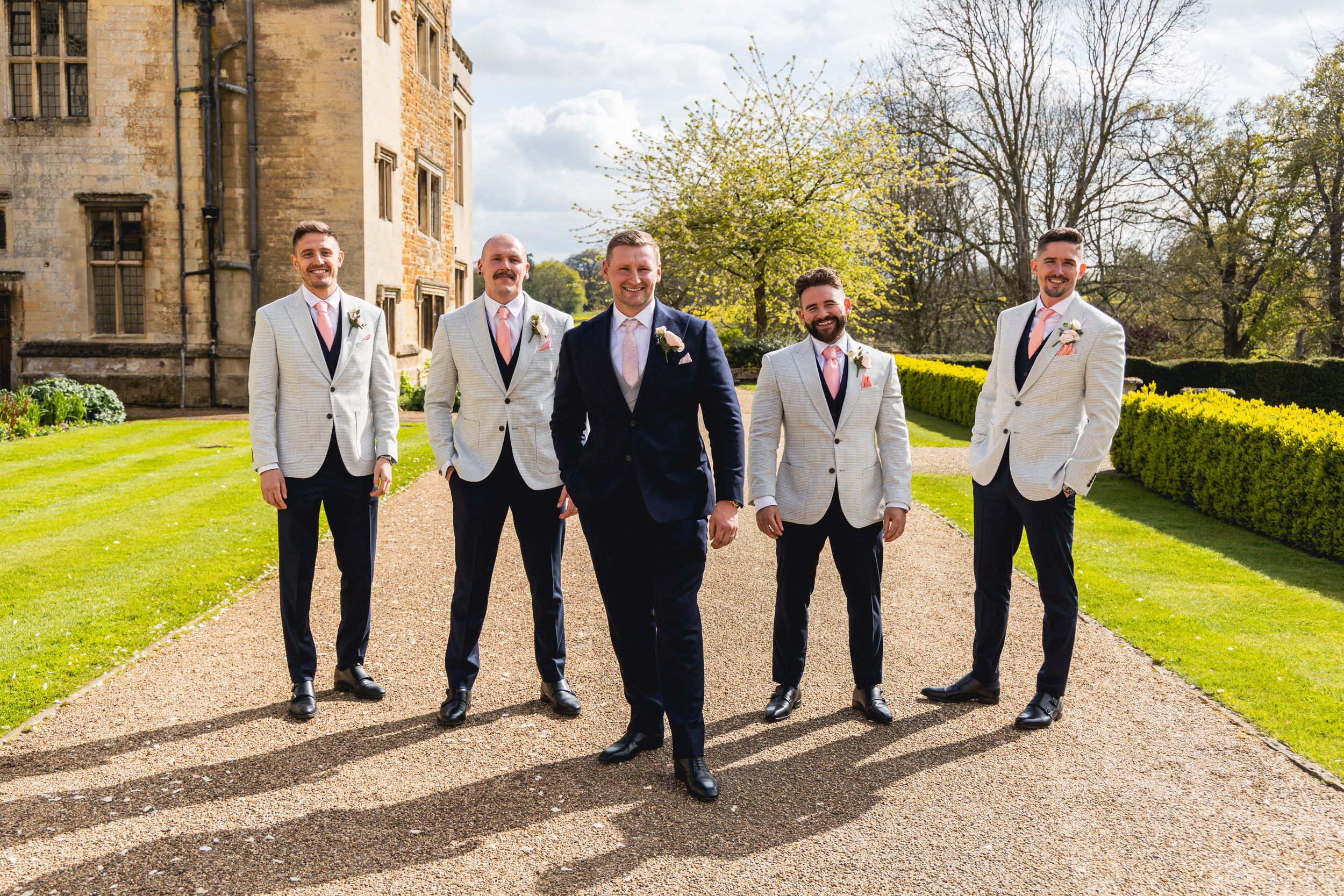 Groomsmen in light gray suits and navy suit pose together on garden grounds.