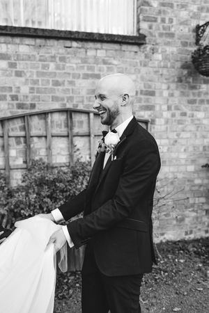 A person in a black suit holds and adjusts a white garment outdoors against a brick wall background.