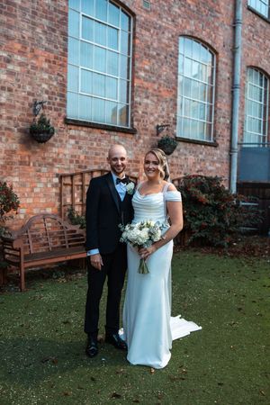 A couple poses together on a grassy lawn outside a rustic brick building with large windows.