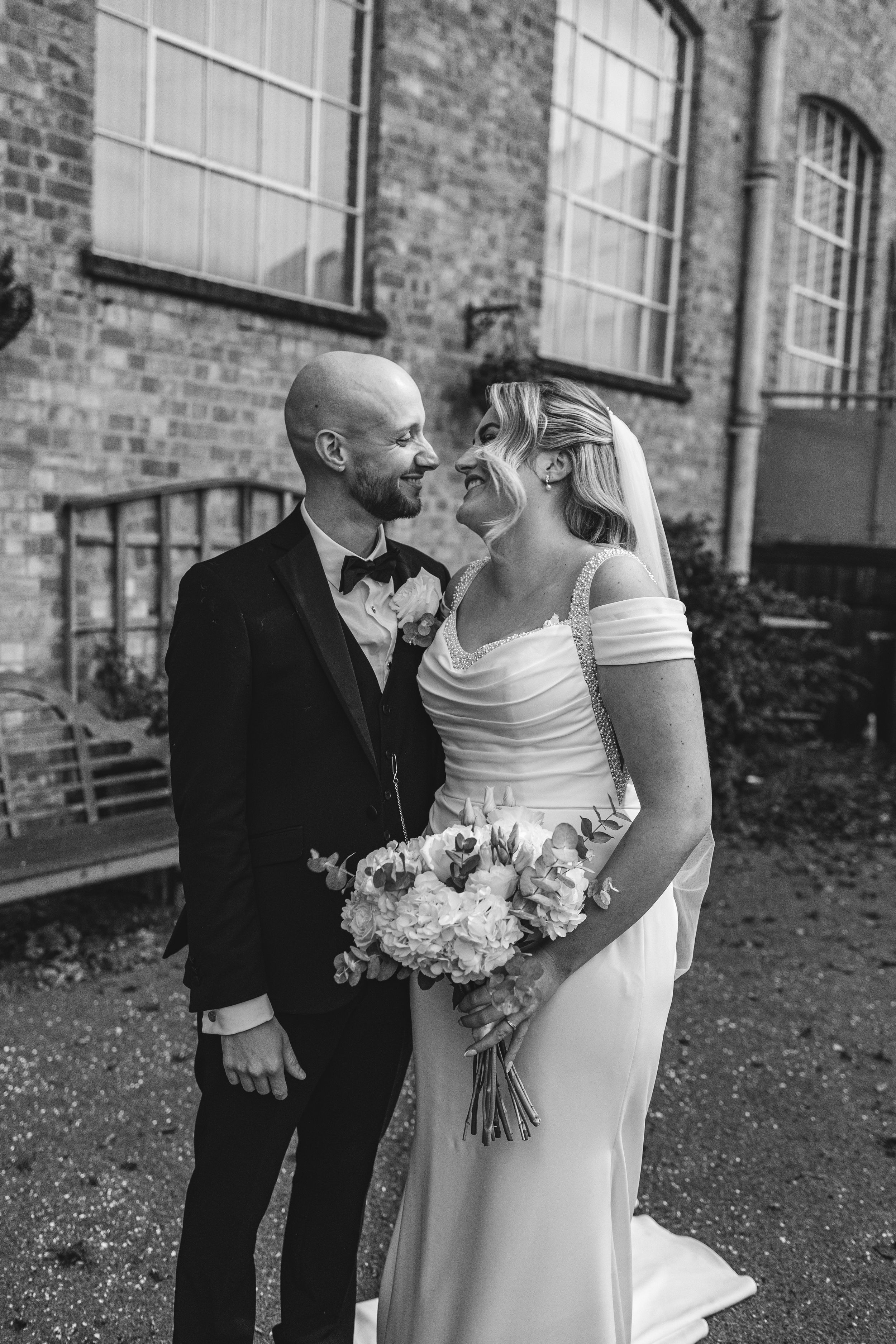 A couple shares a tender moment in formal wedding attire outside a brick building with white flowers.