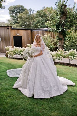 Bride in elegant lace ball gown with cathedral veil holding white bouquet, posing in lush garden setting.