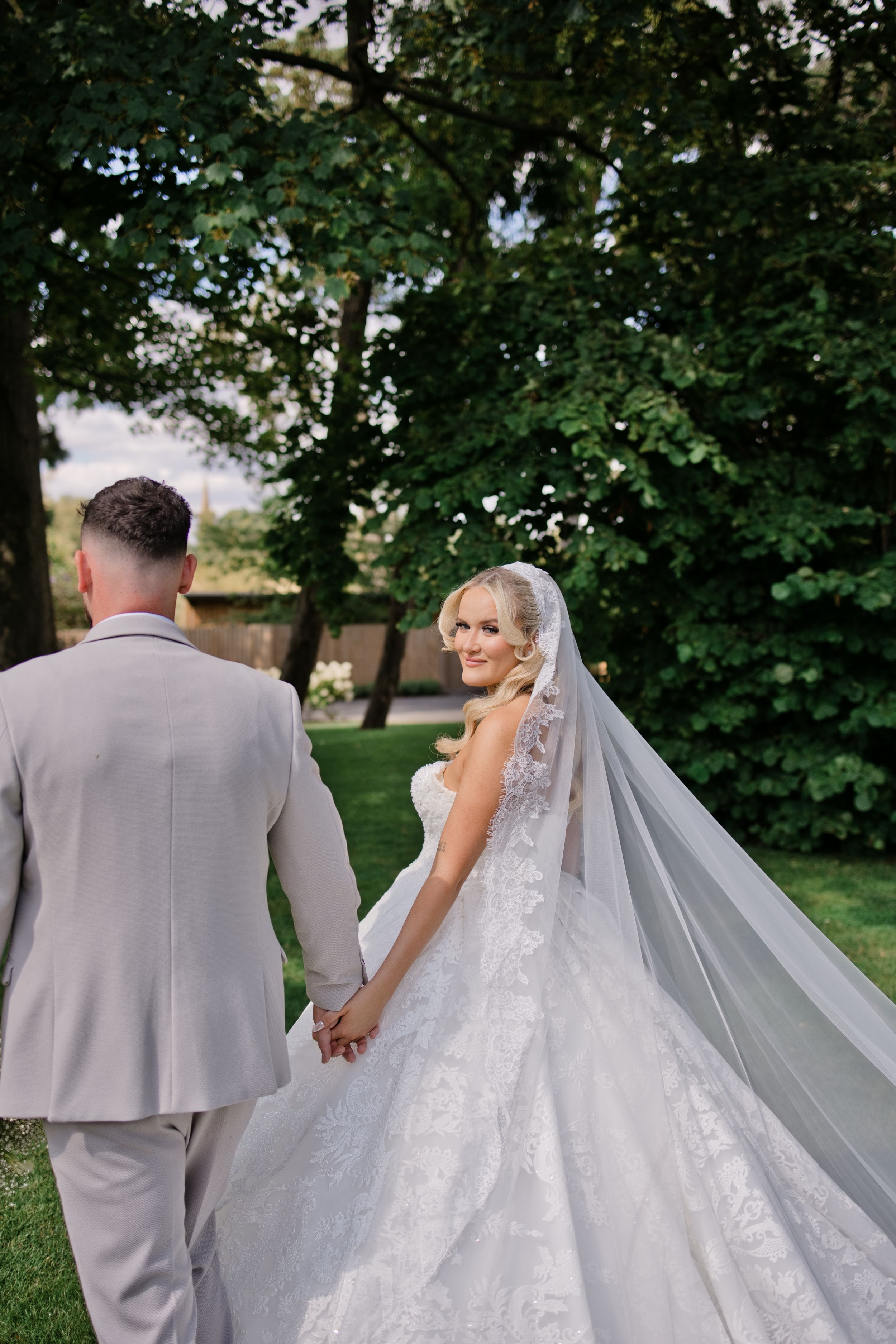 Bride in lace gown and veil holds hands with groom in gray suit while walking through lush garden.
