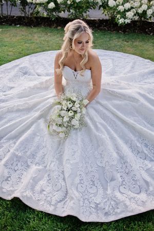 Bride in strapless lace ball gown wedding dress holding white floral bouquet, seated on green grass with full skirt spread.