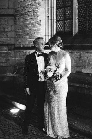 Elegant black and white wedding photo of a couple in formal attire with bridal bouquet standing against stone church wall.