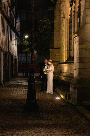 A couple in wedding attire stands illuminated by warm light against a historic stone building on a cobblestone street at night.
