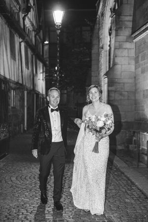Couple walking down a cobblestone street at night, wearing formal attire with bride holding bouquet and groom in tuxedo.