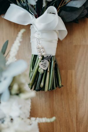 Bridal bouquet with white ribbon, pearl bracelet, and heart charm reading with you every step on wooden floor.