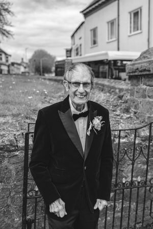 Elderly man in tuxedo with boutonniere smiling at wedding, black and white portrait outdoors.