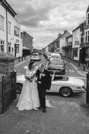Bride and escort walking to a classic white car on a UK high street in black and white photo.
