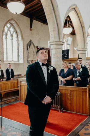 Groom in black tuxedo standing at church altar, looking up during wedding ceremony.