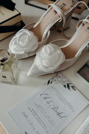 Elegant bridal flat lay featuring nude heels with white rose accents, a wedding invitation for Ed and Rachel, and perfume.