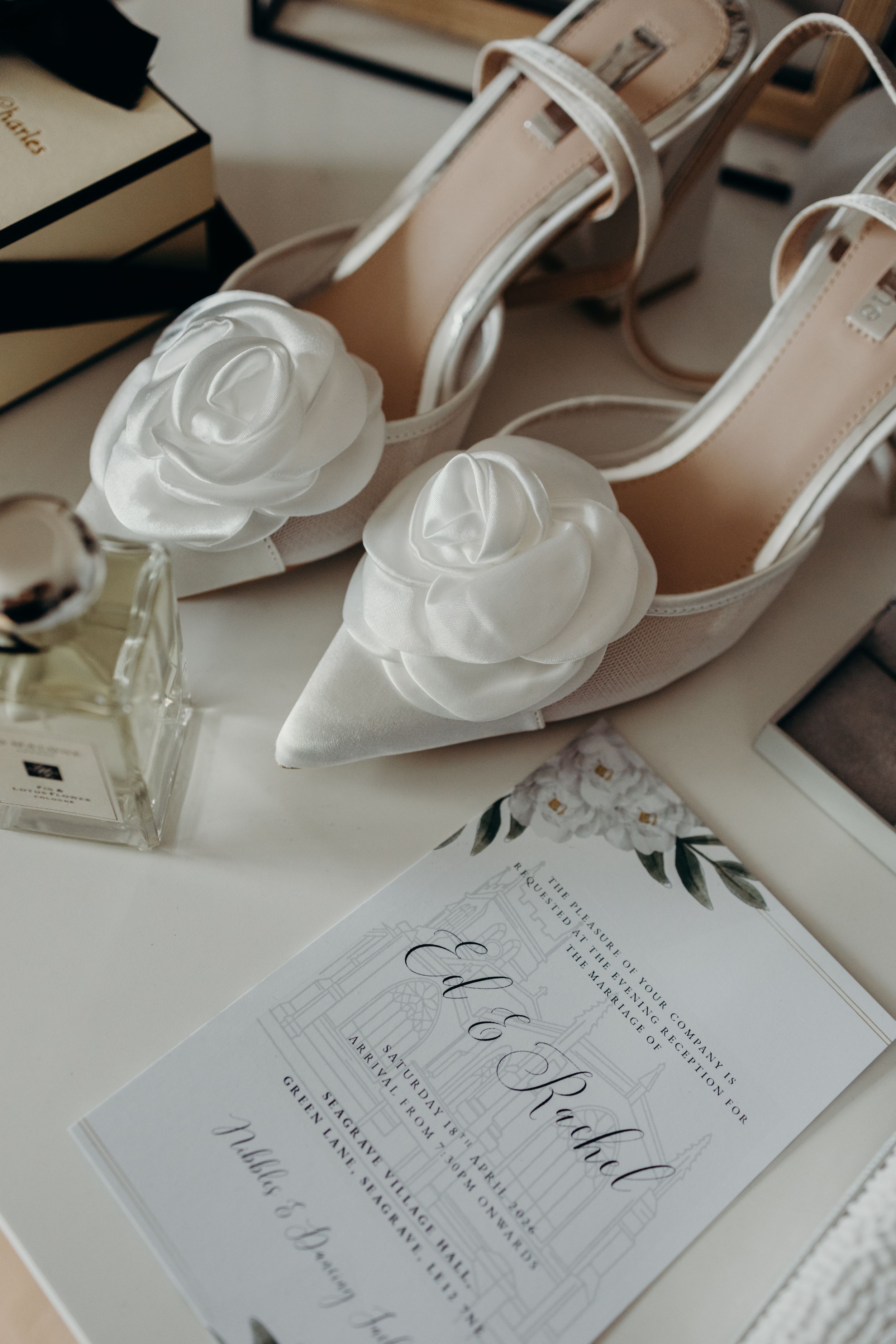 Elegant bridal flat lay featuring nude heels with white rose accents, a wedding invitation for Ed and Rachel, and perfume.