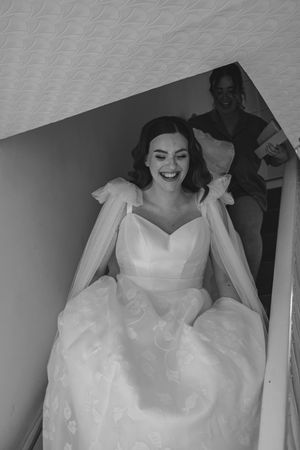 Joyful bride in strapless ball gown with tulle cape laughing while descending staircase in black and white photo.