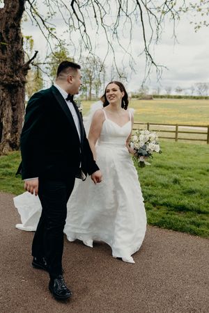 Bride in white gown and groom in black tuxedo walk hand in hand along a garden path on their wedding day.