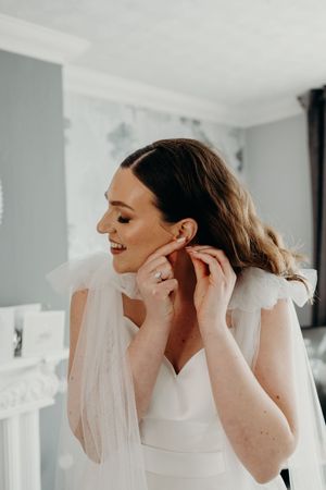 Smiling bride in white dress adjusting her earring while getting ready on her wedding day.