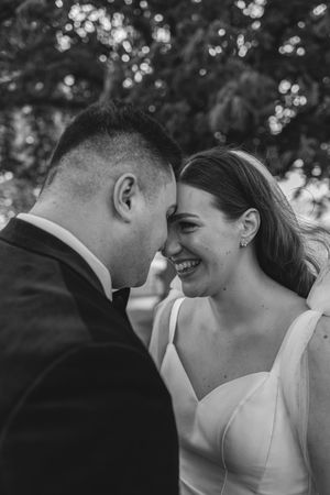 Black and white photo of a laughing bride and groom touching foreheads outdoors.