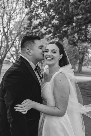 Groom kissing bride on cheek as she smiles joyfully in a white gown and veil in a black and white wedding portrait.