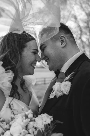 Black and white photo of smiling bride and groom touching foreheads under a flowing veil with a floral bouquet.