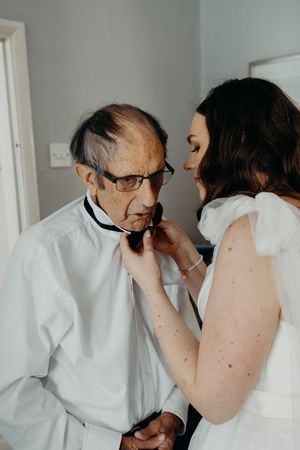Bride helping elderly man adjust black bow tie, both dressed in white for a wedding ceremony.