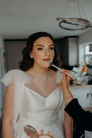 Bride getting makeup applied with a brush while wearing a white wedding dress with flutter sleeves.