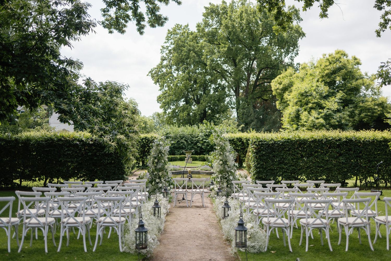 White wedding chairs arranged outdoors in rows for a garden ceremony beneath leafy green trees on a sunny day.