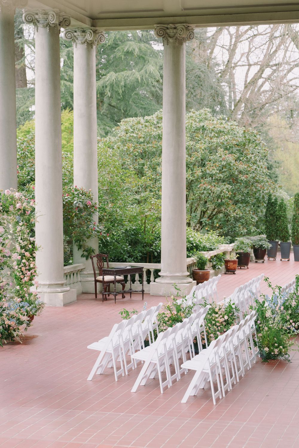 Elegant outdoor wedding ceremony setup with white folding chairs on a brick terrace surrounded by lush gardens and tall white columns.