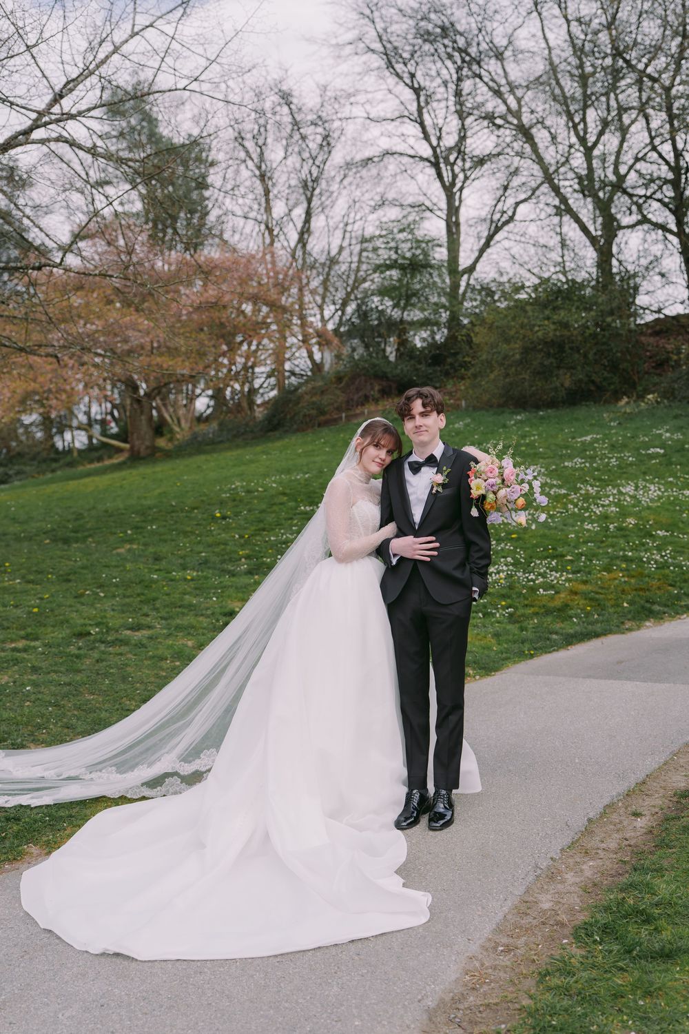 Bride in white ball gown with long veil stands with groom in black tuxedo on garden path surrounded by lush greenery.