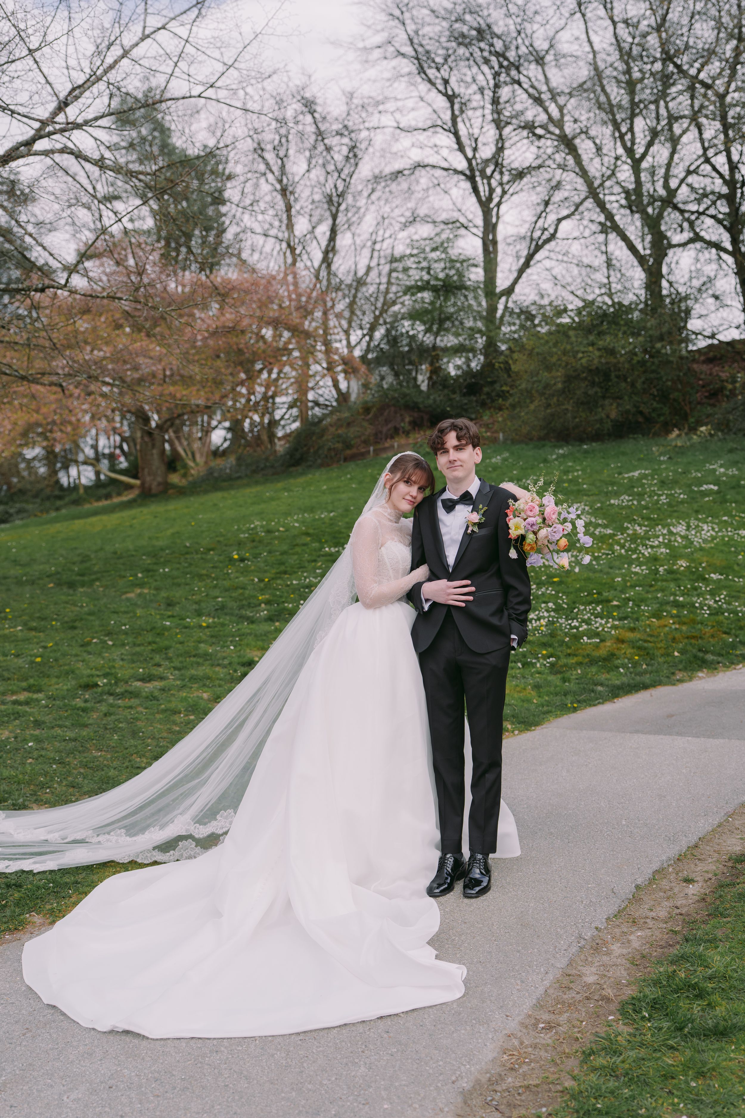 Bride in white ball gown with long veil stands with groom in black tuxedo on garden path surrounded by lush greenery.
