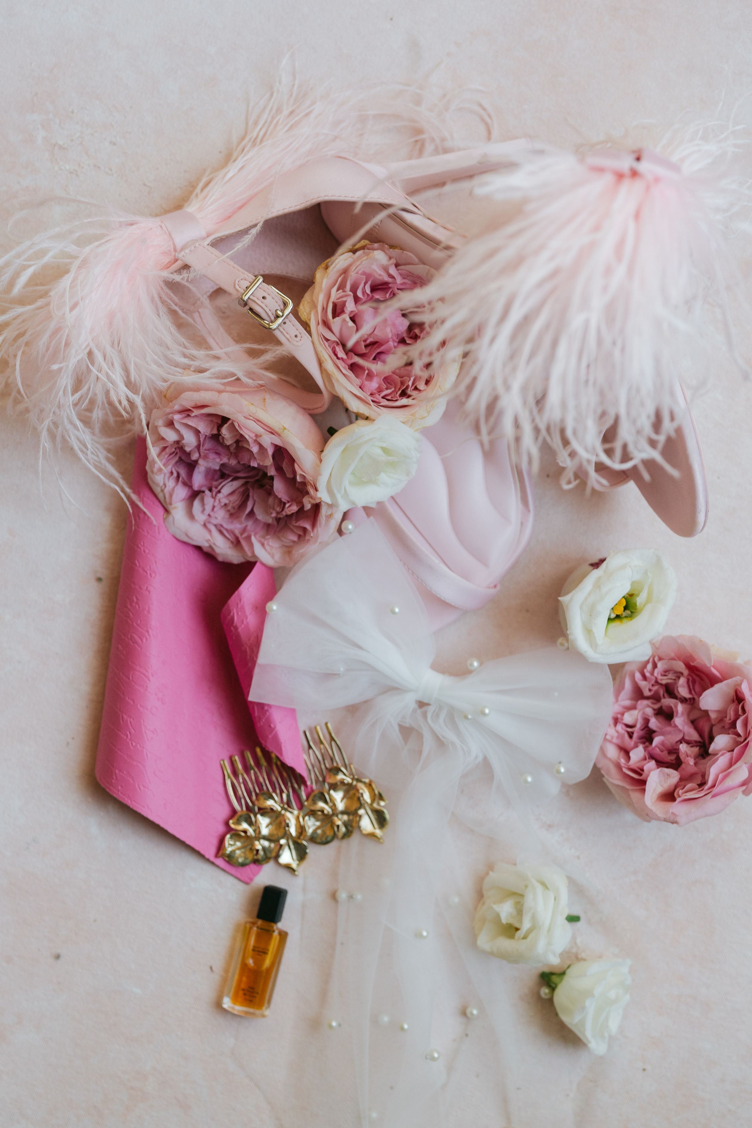 Elegant wedding details featuring pink roses, white feathers, and perfume bottles arranged on a light surface.