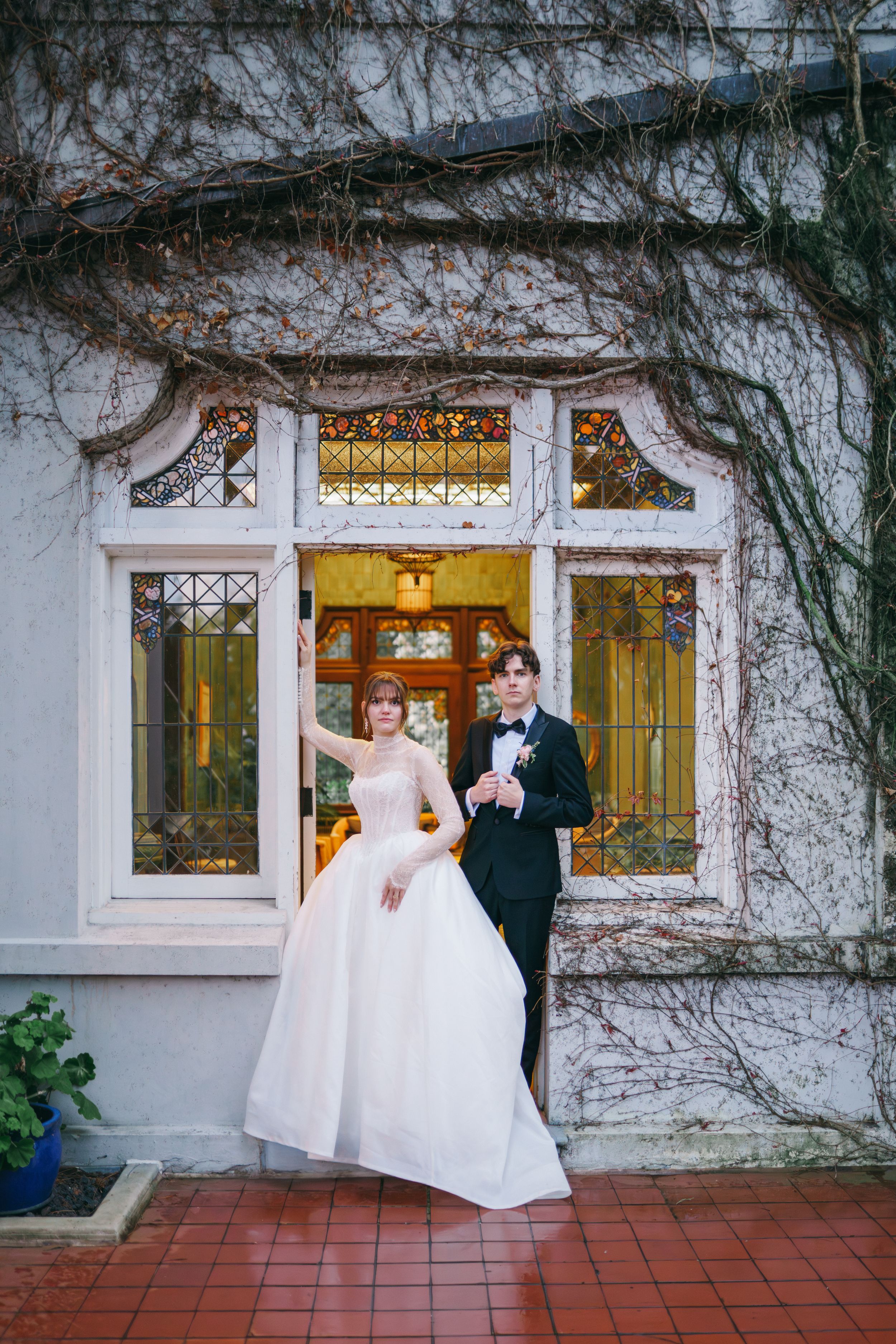 Bride in white ball gown and groom in black tuxedo posing before a vine-covered white building with ornate windows.