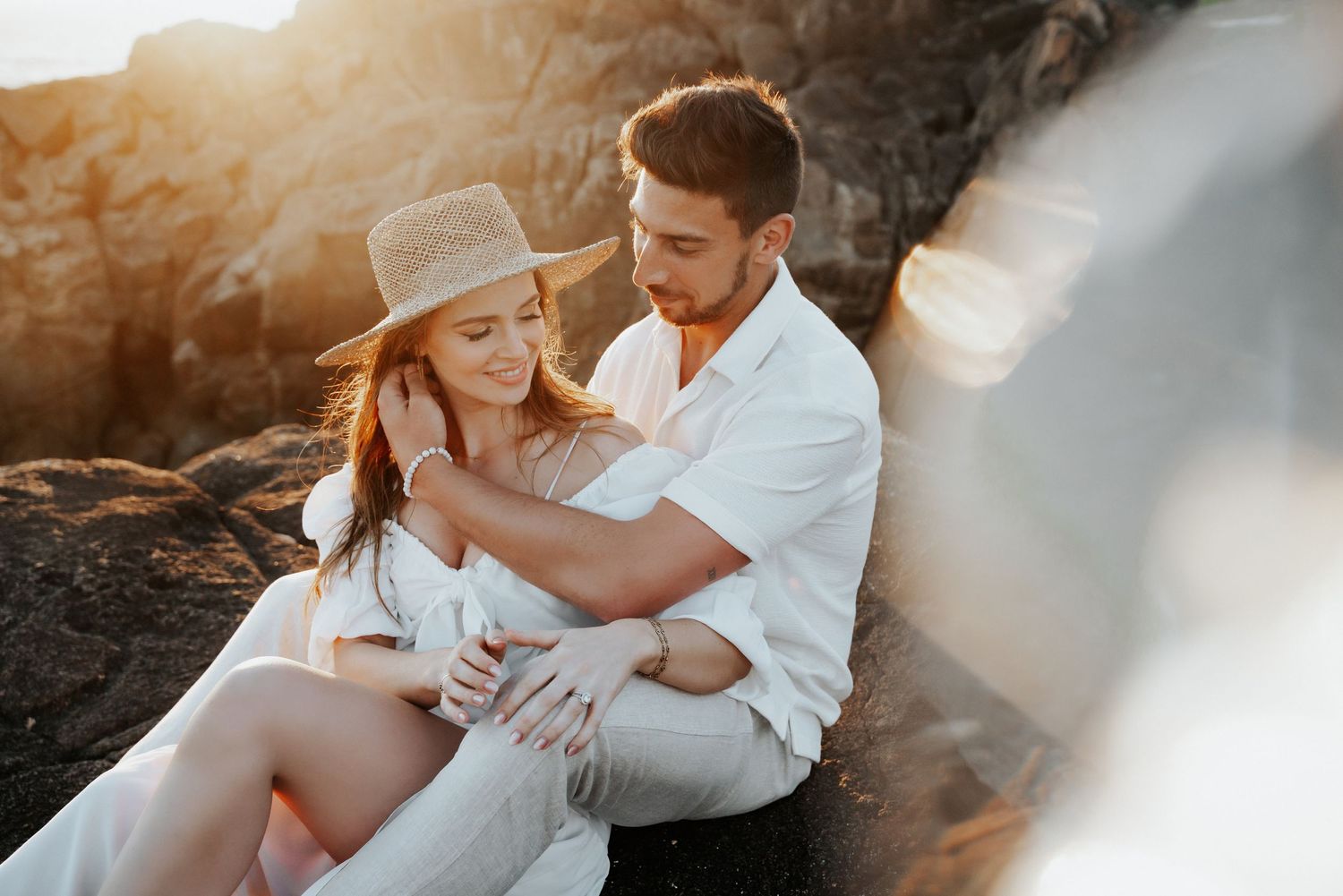 Couple in white outfits share romantic moment on rocky coastline at sunset.