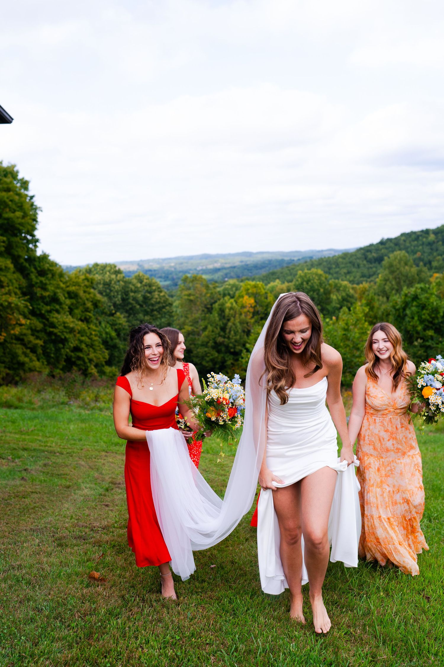 Bride and bridesmaids walking through grassy field in colorful dresses with mountain views.