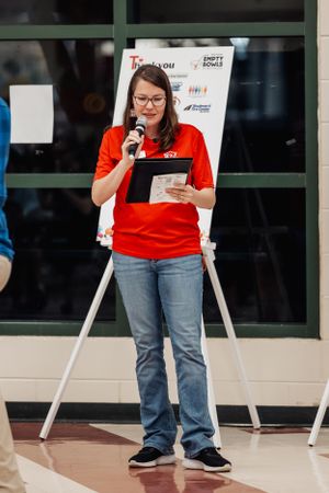 Volunteer in red t-shirt speaking at podium during community event.