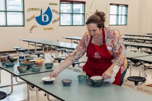 Person in red apron arranging bowls on cafeteria tables.