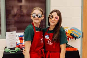 Two people in red aprons and festive glasses posing at community event.