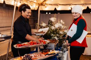 Two people serving food at a catered holiday event under string lights.
