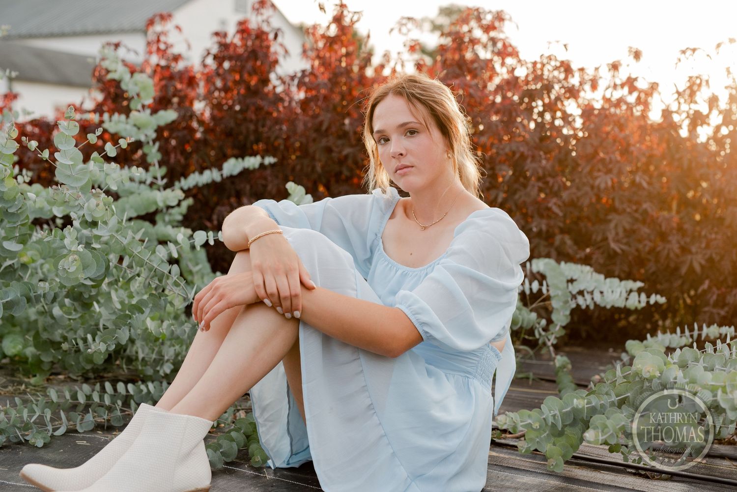 Someone in a pale dress sits outdoors during golden hour with autumn foliage in background.